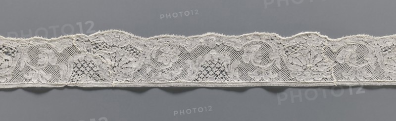 Lace fragment, lace, mesh background with rounded meshes. The lower edge is slightly serrated. The floral motifs on the edge are emphasized with thicker thread. 2nd half of the 18th century, Mechelen. Lace, width 4.2 cm, length 57 cm., A long narrow strip of delicate lace with a scalloped top edge and a straight bottom edge rests against a smooth background; the lace features a repeating floral and scrollwork pattern formed by open mesh areas and more densely stitched motifs, with visible loops and tiny decorative picots along the scalloped edge and a thin stitched border along the bottom; the lace appears off-white or cream against a medium gray background, and there are several areas where the threads form intricate nets and solid embroidered shapes.