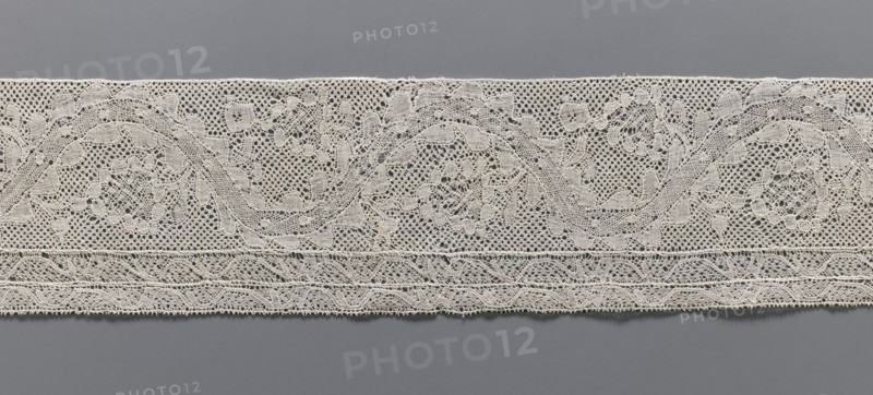 Lace fragment, bobbin lace Malines/Mechelen, 18th century; plant ornaments with floral branches and tendrils in the bends of a ribbon; straight edges, bottom edge with two trails repeating the tendril motif schematic; various mesh backgrounds; side edges folded and hemmed; width 11 cm, length 72.5 cm., A long strip of lace with intricate openwork laid horizontally against a smooth background; the lace appears off-white or cream and shows a repeating pattern of swirling floral and foliate motifs created from fine threads and tiny mesh areas, with a denser decorative band near the lower edge featuring braided and scalloped shapes; the background is a uniform gray that contrasts with the lighter lace, and the overall image emphasizes the texture and delicate holes of the fabric.