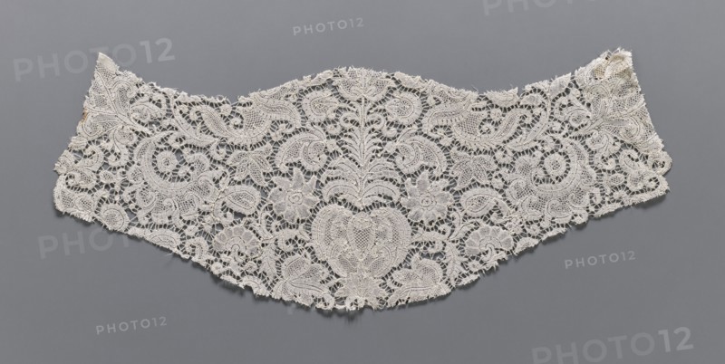 Fragment of lace, plant ornaments, bobbin lace, 18th century, Flanders, width 17 cm, length 46 cm, A delicate lace fabric piece rests flat against a gray background, showing an intricate openwork pattern of floral and foliate motifs with scalloped edges. The lace appears off-white to cream in color, with a few areas of slightly darker discoloration near one edge. The design includes several stylized flowers, curved leaf shapes, and swirling paisley-like forms, all rendered in fine thread with textured raised outlines and mesh-filled interiors. The fabric has a slightly irregular outer contour with frayed thread ends and tiny gaps where the lacework is broken, and the overall appearance is light, airy, and detailed against the solid gray surface.