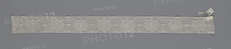 Fragment of lace, bobbin lace, 18th century, Flanders, lace, width 9 cm, length 51 cm, A long narrow strip of delicate lace laid flat against a grey background, showing an openwork pattern with a repeating floral motif and a fine net-like ground; the lace is off-white to cream in color, with subtle variations in tone along its length; at one end there are a few small attached tags and a short length of green thread visible.