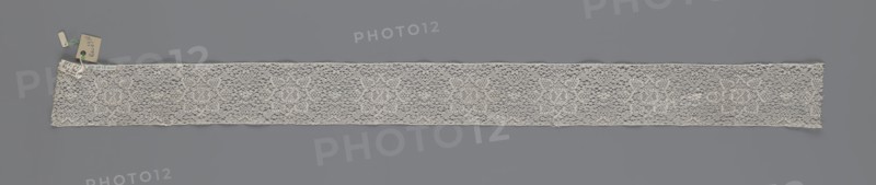 Fragment of bobbin lace, 18th century, Flanders; width 9 cm, length 51 cm; subject: lace; maker, provenance includes repossession; place: Henryków German: Heinrichau., A long narrow strip of openwork lace laid flat against a solid muted gray background, the lace appearing off-white to cream with a delicate, airy texture and an intricate repeating floral-like motif visible across its length, surrounded by a fine netted ground; one end of the lace has a small cluster of tags attached with thin strings in pale beige and light green hues, and the lace edges are slightly irregular with subtle scalloping and fine threadwork.