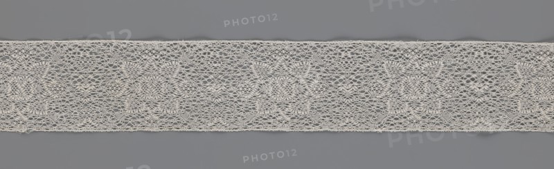 Fragment of bobbin lace, 18th century, Flanders, lace, width 9 cm, length 51 cm, A horizontal strip of delicate lace with an open, airy weave stretches across a gray background; the lace is an off-white or cream color with an intricate repeating pattern that includes round floral motifs and scalloped edges, and the background shows varying shades of medium gray that contrast with the lighter lace.
