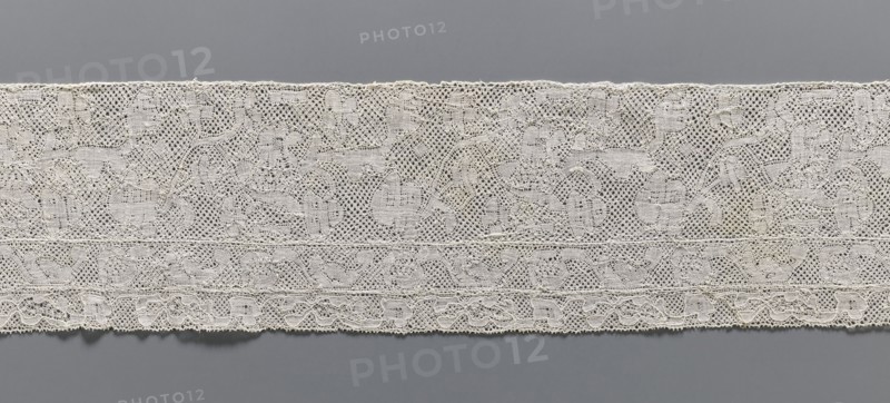 Lace fragment, bobbin lace Binche lace, 18th century. Background: mesh with large, rounded meshes. Edges of motifs uncontoured. Large, narrow laces are sewn on at the bottom edge; the wider one has a motif of flower branches arranged in a twig; the other, narrower one, has a motif of a flower tendril. Width 12 cm, length 61 cm., A horizontal strip of lace displayed against a solid background, showing a delicate openwork pattern with a floral and scroll motif. The lace appears in shades of off-white and cream with subtle variations in tone, and the background behind and below the lace is a smooth gray. The lace has a scalloped lower edge and a slightly uneven upper edge, with a few rows of denser woven bands running parallel to the edges. The overall texture is intricate and airy, with small holes and looping threads forming repeating decorative shapes.