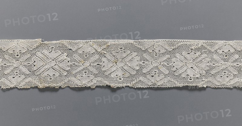 Lace fragment, bobbin lace with rosette ornaments framed by a tendril motif at top and bottom; mesh background similar to a drochel; motif edges contoured and straight. 18th–19th century, Flanders. Lace, width 8 cm, length 97.5 cm., A horizontal strip of lace with a repeating geometric floral pattern spans the width of the image, placed against a smooth background. The lace appears off-white to cream with small areas of darker discoloration, and the background shows shades of gray ranging from medium to darker gray. The lace has an open, net-like texture with solid embroidered motifs forming diamond and cross shapes, and the lower edge is slightly irregular with tiny loose threads. The overall scene shows the contrast between the delicate light-colored lace and the uniform gray backdrop.