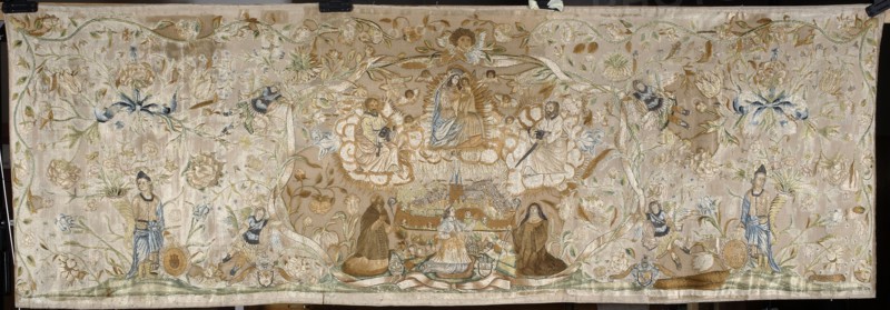 Antependium by Anna Barbara Orliec embroiderer, 1693. Antependium embroidered with colorful silks on a creamy pink satin background, details worked with metal thread; techniques include flat, split, and knotted stitches and metal-braided threads laid flat. Size: height 90 cm, width 274 cm. In the center, within a wide ribbon wreath supported by four archangels in short armor holding palm leaves, is a walled city surmounted by Mary and Child, St. Peter, and St. Paul. Kneeling before the city are a bishop bearing the Junosza coat of arms three lambs, an abbot with the Brinken coat of arms three roses behind a shield resembling crossed crosiers, and an abbess with the Nowina coat of arms. Around them are flowers, a floral tendril, and six angel heads encircling Mary. On the sides are two figures in antique-style robes holding horns filled with flowers and heraldic shields — one possibly showing the coat of arms of Kraków a city gate with an eagle, the other illegible. At the bottom are tw...