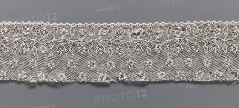 Lace fragment, needle lace Alençon lace, A flower branch parallel to the edge. Above the branch, in the projection, circles made of small circles. Below the branch, on the edge, a row of small circles, the edge corresponding to the circles—small semicircular serrations. 18th century, France, lace, width 6.5 cm, length 32 cm, A horizontal strip of delicate lace in shades of off-white and cream lies against a flat gray background, showing a repeating floral and geometric pattern with a scalloped top edge and a straighter bottom edge; the lace fabric has a fine hexagonal mesh with a few raised knot-like floral motifs and looping vine-like designs, and there are small irregular frayed threads along the lower edge.