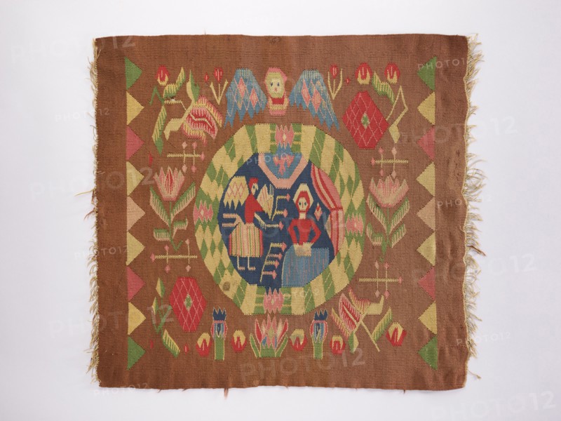 Ornamental-figural tapestry, flowers, floral ornaments, angels, Copernicus, Mary mother of Jesus Christ, Annunciation Bible; New Testament, furniture upholstery, interior furnishings, weaving, tapestries. In the central medallion are the Virgin Mary and an angel. Outside the medallion are flower branches, heads, and other decorations. The ornament is strongly stylized and geometric. Without a border. Colors: light brown, navy blue, red, yellow, blue, and green. 18th century, Norway. Fabrics; height 59 cm, width 62 cm., A rectangular woven textile with fringed short edges displays a central circular medallion with a dark blue interior containing stylized human figures and geometric shapes, surrounded by a green and pale yellow checker border; the field around the medallion is a warm brown and features symmetrical floral and geometric motifs in red, pink, green, pale yellow, and blue, with a repeating triangular border pattern along the left and right edges and frayed threads visible at ...