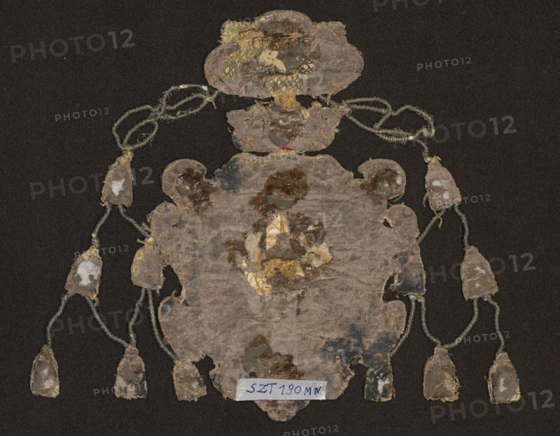 Coat of arms, embroidery, provenance message: Vasas family; coats of arms: Vasa sheaf on central shield. In four fields: 1 Eagle with a crown — Polish; 2 Three crowns — Swedish; 3 Pogon — Lithuanian; 4 Gryf — Ruthenian. Shield topped with a crown and a bishop's hat. Possible owners: John Albert Snopek coat of arms, Bishop of Kraków, cardinal, died 1634; Jan Kazimierz Snopek coat of arms, elected King of Poland 1648, abdicated 1668, titular cardinal; or Ferdynand Vasa, brother of Jan Kazimierz. Embroidery background: linen canvas with silk satin in red and blue applied to canvas. Embroidery on a backing of various heights made of linen threads. Flat and convex embroidery with metal threads: silver and gold plate braided on silk yarn; drawn wire in silver and gold; bullions of drawn wire and narrow gold plate. Silk embroidery in satin and knot stitch. Attributed to E. Soszko; ca. 17th century, Poland. Materials: fabrics; height 16 cm., An irregularly shaped, worn textile or paper object ...