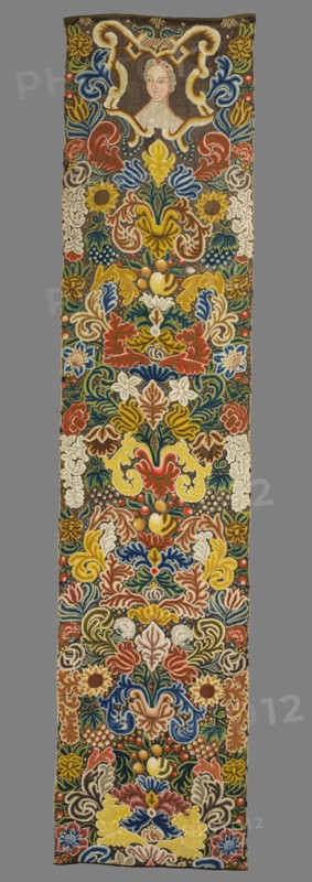Tapestry. Tapestry embroidered in wool and silk on linen canvas. In the upper part, in a decorative cartouche, is the bust of a woman with two red flowers pinned in her hair. Around it are dense scrolls of large leaves, flowers, and fruit. Unedged. Vivid colors: brown background, red, pink, green, sapphire blue, yellow, olive, and ochre ornament. The plant motifs of the nine fabrics comprising this set do not differ from each other. Stylized leaves, flowers, and fruit fill the entire surface of the panel. The composition unfolds on both sides of the axis, defining a symmetrical division of the decoration, similar to the large-pattern silk upholstery fabrics fashionable in the late 17th and early 18th centuries. Minor differences include the inclusion of small animal silhouettes on some panels, which enrich the decoration. All fabrics are decorated with the same fruit: plump bunches of black and white grapes, yellow apples, and dark red cherries. Vivid colors, brown background, shaded o...