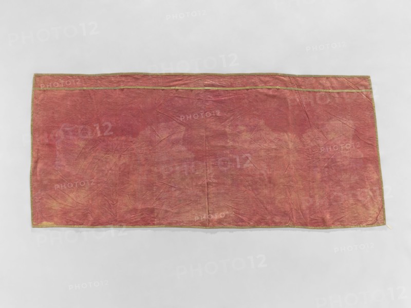Church napkin, gift, church paraments, evangelical churches furnishings, Sielec Swietokrzyskie Voivodeship, Staszów County, evangelical church Sielec - furnishings; motif: flowers, leaves, plant ornaments; sacred art; churches and chapels furnishings; date: 1700–1730; origin: France; material: fabrics; dimensions: height 97 cm, width 216 cm, depth 10 cm., A rectangular fabric laid flat against a light gray background, primarily a faded reddish color with areas of muted pink, peach, and tan showing through; the fabric has visible creases and a slightly wrinkled texture, and a narrow decorative trim in a goldish color runs along the edges.