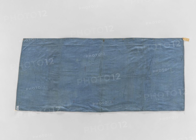 Church napkin; creator, ; church paraments; evangelical churches furnishings; Sielec Swietokrzyskie Voivodeship, Staszów County; evangelical church Sielec — furnishings; decoration: flowers, leaves, plant ornaments; sacred art; churches and chapels furnishings; dated 1700–1730; origin: France; material: fabric; dimensions: height 97 cm, width 216 cm, depth 10 cm., A rectangular piece of fabric laid flat against a light gray background, appearing blue with variations in shade and subtle darker and lighter patches, showing visible creases and folds across its surface, with a hemmed edge and a group of small round metal eyelets along the top edge, a few tiny darker spots and minor imperfections scattered on the cloth, and small fabric tabs at the corners.