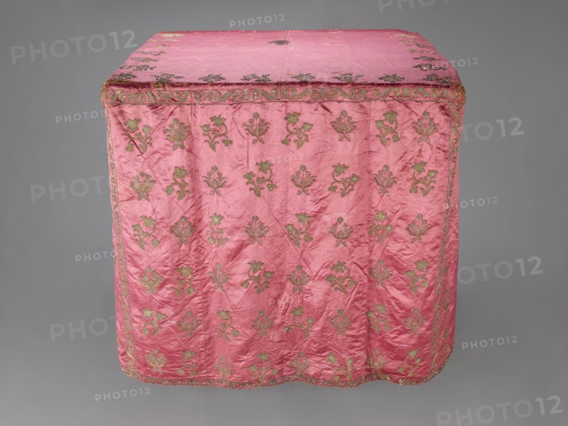 Church cope, liturgical vestment, 17th century, Poland. Made of raspberry satin, embroidered with floral patterns. On the front, branch patterns are arranged in six rows: two branch types—asymmetrical, slightly inclined branches with three flowers, and symmetrical, straight branches with a single flower at the top and two pairs of leaves. Edges decorated with a border of floral and fruit tendrils; lower edges trimmed with gold bobbin lace. Beige linen lining. Height 298 cm, width 201 cm., A rectangular textile draped over a blocky form, predominantly pink with a sheen, featuring a repeating ornate floral motif in a metallic gold-green color across its surface and along its border; the fabric shows visible creases and slight irregularities in texture, with the top panel and sides decorated in the same pattern, and the backdrop is a neutral gray.