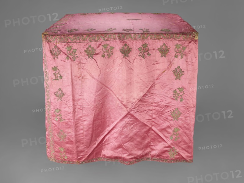 Church cope, gift; liturgical vestment, 17th century, Poland. Made of raspberry satin embroidered with floral patterns. On the front, branch patterns are arranged in six rows: two types — asymmetrical, slightly inclined branches with three flowers, and symmetrical, straight branches with a single flower at the top and two pairs of leaves. Edges decorated with a border of floral and fruit tendril motif; lower edges trimmed with gold bobbin lace. Beige linen lining. Height 298 cm, width 201 cm., A draped pink fabric panel with a slightly wrinkled texture covers a rectangular form, showing a decorative border and repeating ornate motifs embroidered near the edges; the embroidery appears in a metallic gold-green tone contrasting with the pink, and the background around the panel is a neutral gray.