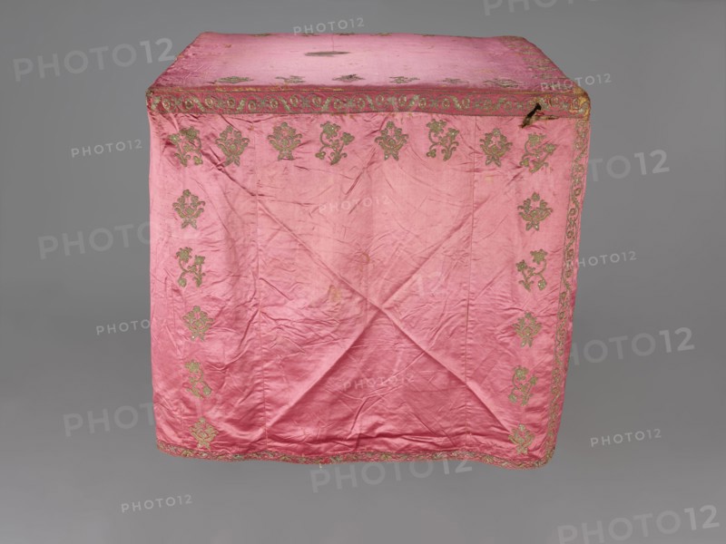 Church cope, liturgical vestment, 17th century, Poland. Made of raspberry satin, embroidered with floral patterns. On the front, branch patterns are arranged in six rows: two types—asymmetrical, slightly inclined branches with three flowers, and symmetrical, straight branches with a single flower at the top and two pairs of leaves. Edges decorated with a border featuring a floral and fruit tendril motif; lower edges trimmed with gold bobbin lace. Beige linen lining. Height 298 cm, width 201 cm., A large pink fabric draped over a rectangular form with visible creases and folds, decorated with a repeating ornate floral motif in a greenish-gold color along the top edge and down the sides, and a decorative border in the same greenish-gold color running around the top and bottom edges; the fabric shows areas of wear and small dark spots, and the background is a uniform gray.