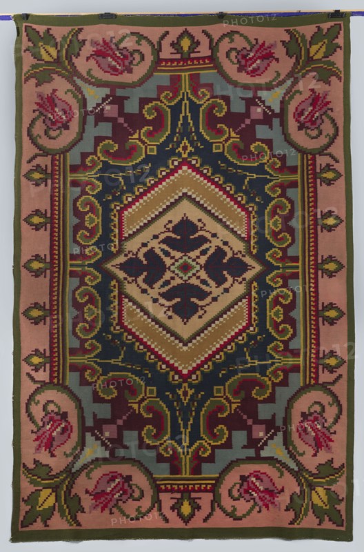 Kilim, decorative flatwoven textile, 19th/20th century, Kosów; manor and house furnishing; decorative fabric; height 304 cm, width 202 cm., A rectangular woven textile with a central diamond-shaped medallion surrounded by ornate geometric and floral motifs in a palette of deep navy, olive green, muted pink, mustard, burgundy, beige, and pale blue-gray. The central area features layered chevron bands in beige and mustard framing a dark navy inner diamond with symmetrical stylized leaf shapes in burgundy and navy. Around the medallion are scrolling arabesque forms and stepped geometric blocks in olive, mustard, burgundy, navy, and blue-gray. The border consists of a muted pink field with repeating roundels containing stylized floral forms in shades of pink and burgundy, linked by olive green foliage and framed by narrow bands of olive and mustard. The overall appearance is richly patterned with pixelated, tapestry-like stitching and contrasting color edges.