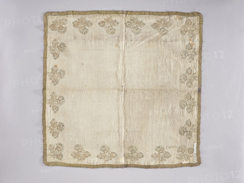 Velum, embroidery, church paraments, floral motifs, Henryków Heinrichau. Flowers, leaves, plant ornaments; sacred art; churches and chapels furnishings. Velum or church cloth of white linen batiste, embroidered along the edges with floral bouquet motifs in gold thread. In the center is an embroidered inscription IESUS, above which a small cross. Around the edges are stylized Persian-style flower bouquets. In one corner are embroidered the letters HHGS. Trimmed with gold bobbin lace with an interwoven ribbon motif late 17th century. A wide approx. 3 mm strip of gold thread is sewn onto the edges where the lace is sewn. Double-sided embroidery. Flat, pierced embroidery, satin stitch. The stitch direction is adapted to the shapes of the ornaments. Metal threads — a gold plate covered with multicolored patina, woven on a silk base. 17th century, Silesia; liturgical vestment. Height 74 cm, width 82 cm., A square cloth with a pale cream background bordered by a textured gold trim, showing a ...