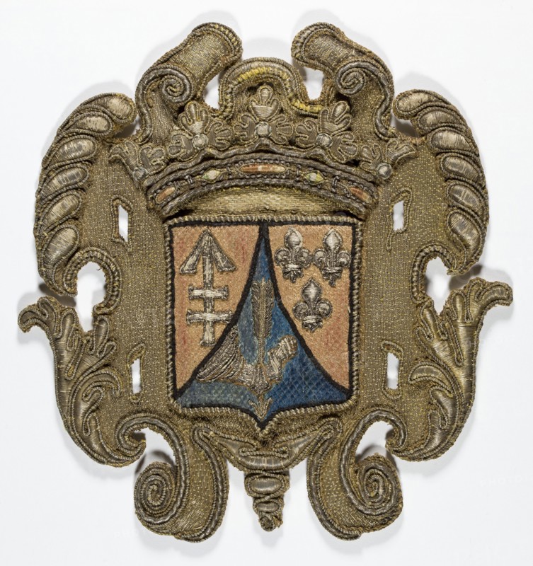 Coat of arms cartouche, embroidery; coat of arms cartouche with a tripartite shield under a crown in the center. Coats of arms: Lis, Kikieryc, unknown most likely the coat of arms of the Sapieha family. 17th century, Poland. Materials: fabrics. Dimensions: height 22 cm, width 21 cm. ., An ornate embroidered shield-shaped patch with an elaborate scrollwork border in metallic gold tones; the central shield area is divided with a triangular blue section and surrounding tan sections, and contains embroidered motifs including a winged element in blue and tan and three fleur-like shapes in metallic tones; the top of the patch features a crown-like decorative element in gold, and the entire piece shows textured stitching, raised metallic threads, and areas of darker and lighter gold, along with visible tan, blue, and muted silver-gray hues.