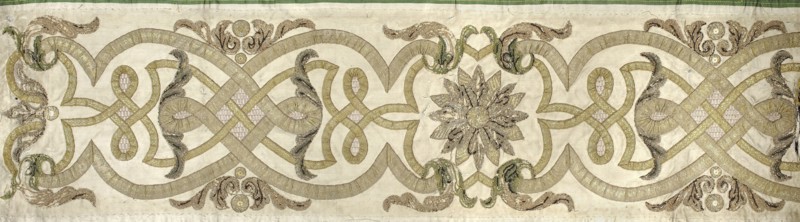 Fragment of a chasuble column, embroidery, rosettes, ribbon-and-pincer ornament. The column from the back of the chasuble is made of cream satin, onto which narrower and wider gold braids are sewn, creating a pattern of interwoven ribbons. A rosette and acanthus leaves are placed among them. The rosettes and leaves are embroidered with gold thread, sequins, and a slit. Early 18th century, Poland; fabrics; length 95 cm, width 26 cm., A horizontal decorative textile panel with a pale beige background featuring an intricate continuous knotwork pattern in shades of gold and muted olive, punctuated by a central floral rosette composed of layered petal shapes in grayish gold and darker brown tones; the design includes several looping bands and ribbon-like elements in gold with subtle pale pink textured inserts, flanked by symmetrical scrolling foliage motifs in green, brown, and gold, and accented by a few circular medallion shapes in golden and brown hues, with a narrow green border along t...