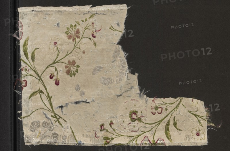 Fragment of white silk grosgrain brocaded with colorful threads and silver stitching; brocade threads include silver stitching, silver metal thread, light celadon, green, grass green, burgundy, apricot, dark pink. Grosgrain background brocaded with small twigs with twining shoots and fancy flowers; drawing includes a rhomboidal mesh net. 18th century, France. Fabric fragment, height 31 cm, width 23.9 cm., A worn rectangular fabric fragment with frayed edges sits against a dark background, showing a faded cream base with a floral embroidered pattern in green, pink, burgundy, and pale brown, including stems, leaves, and blossoms; areas of the fabric have small tears and worn spots revealing lighter and slightly bluish threads, and there are a few scattered darker smudges and thin loose fibers along the edges.