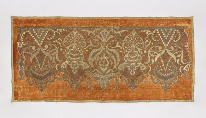 Antependium, antependium; dark yellow velvet with appliquéd decoration of stylized palmettes and flowers in vases in the Louis XIV style; appliqué of gold lace and fragments of gold lamé; edges trimmed with gold galloon; 18th–19th century, Poland; fabric; height 102 cm, width 222 cm., A rectangular textile with a burnt orange background framed by a narrow pale gold border, featuring an ornate symmetrical embroidered band across the upper portion in muted gold and gray tones; the embroidery forms a repeating pattern of stylized floral and foliate motifs with curved, scalloped and lacy shapes, bulbous hanging elements, and flowing tendrils that create mirrored pairs and central vertical designs, with areas of fine mesh-like stitching and solid filled shapes; the overall palette shows burnt orange, pale gold, gray, and subtle brown variations, and the fabric displays slight variations in texture and sheen.