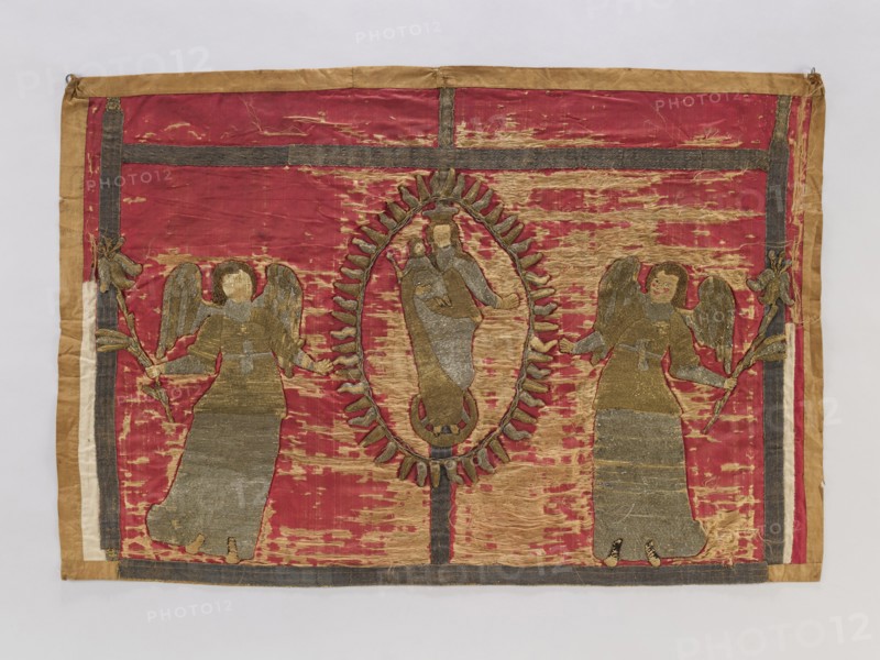 Antependium, antependium;, Raised embroidery applied to red silk satin: in the center Mary and Child in radiant glory on a crescent; at the sides two angels holding lilies. Trims of silver; 17th-century galloon. Embroidery on linen backings of varied heights, applied to silk satin; silk thread split stitch for body parts, metal thread laid stitch for other elements. Metal threads in gold and silver: cut plate, plate woven on linen base, drawn wire. 17th century, Poland. Fabric; height 85 cm, width 126 cm., A rectangular textile with a worn deep red background bordered by tan and dark gray bands, featuring a central oval medallion outlined in tan and dark gray with a figure inside rendered in faded gold and gray tones, flanked on either side by a pair of standing winged figures made of similar gold and gray stitched fabric holding stylized branches, all showing areas of fraying and threadbare wear that reveal lighter tan patches and underlying fibers.