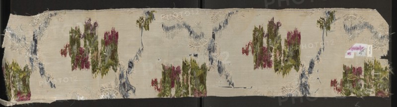 Fragment of fabric. Fabrics, fragments of silk fabrics, patterned fabrics, fabrics with preserved selvedge. Weave: background - Gros de Tour; liseré - white; pattern - brocaded with colored threads and the liseré effect with a white thread creates a subreport. Brocade threads: white, light and dark green, pink, sky blue, pale red, orange, plum. Pattern: floral bouquets connected with lace and chenille ribbons. Report: 43 x ? Edge width: 0.5 cm. France, Lyon probably 19th century; possibly produced in a Lyon manufactory 1820–1840. Height 20.5 cm, width 96 cm., A long rectangular piece of fabric with a light beige background shows a worn embroidered pattern featuring several vertical clusters of frayed threads in shades of green and pink, with accents of reddish tones, scattered across the surface; sinuous vertical and diagonal bands of frayed blue-gray thread run between the clusters, and there are a few areas of pale cream raised embroidery that form partial circular motifs; the fabric...