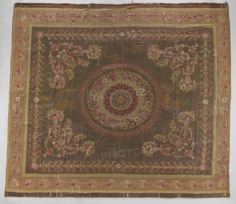 Ornamental tapestry, Late 18th century, Flanders. This ceiling floor tapestry has symmetrical decoration: a central rosette surrounded by two floral and leafy wreaths; corner compositions of horns filled with flowers, lyres, and intertwined acanthus leaves; overall frame of leaves and rosettes. Border repeats leaves, rosettes, and flowers. Colors: cream, olive, and sand background with pink, green, and blue ornament. Materials: woven fabrics. Dimensions: height 517 cm, width 460 cm., A large rectangular textile with a central circular medallion surrounded by floral motifs and ornate corner decorations, featuring a background of muted brown and ochre tones with accents of pink, rose, green, gold, and cream; a broad border in a lighter tan or beige frames the piece, decorated with repeated floral garlands in pink and green, and inner narrow bands in gold; the central medallion has concentric floral patterns in rose, pink, cream, and green, and is encircled by a wreath-like design in redd...