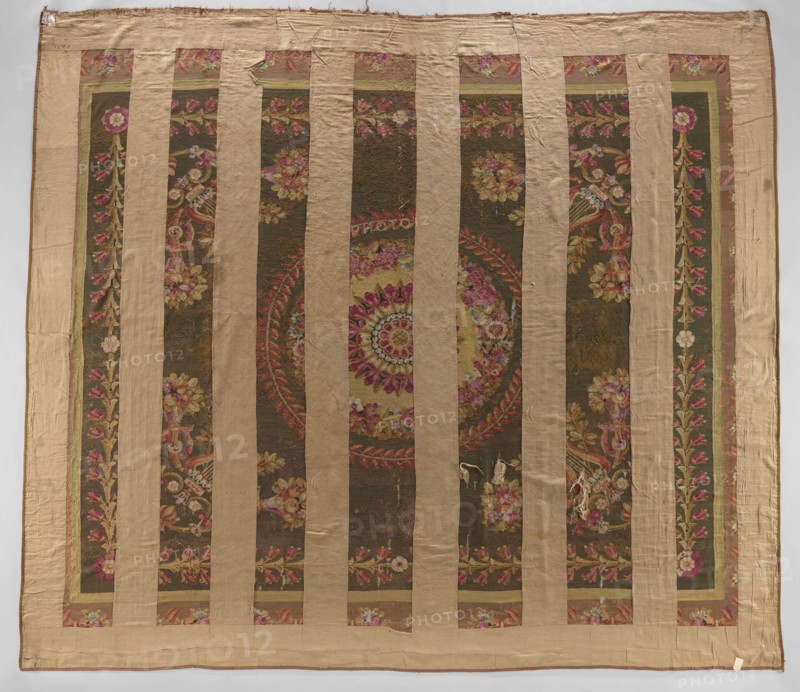Ornamental tapestry, This late 18th-century Flemish tapestry height 517 cm, width 460 cm — cream, olive, and sand ground with pink, green, and blue ornament — features a symmetrical design: a central rosette surrounded by two floral and leafy wreaths; corner compositions of horns filled with flowers, lyres, and intertwined acanthus leaves; an overall frame of leaves and rosettes; and a border with similar motifs. Materials: woven decorative fabric tapestry., A large square textile with alternating vertical bands of plain tan fabric and ornate patterned panels; the patterned panels show a dark olive background with floral and foliate motifs in shades of pink, rose, red, yellow, green, and cream, including a central circular medallion composed of concentric floral rings; the tan bands and border are a muted beige, and there are visible areas of wear and small holes in some patterned sections; the overall colors visible are tan, olive, pink, rose, red, yellow, green, and cream.