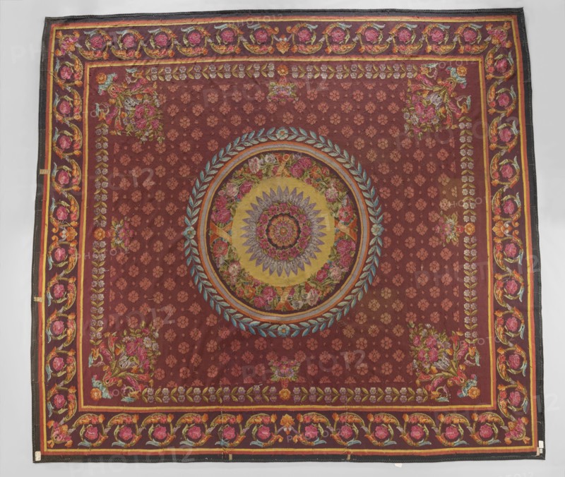 Ornamental tapestry, unknown origin, flowers, floral ornaments, weaving, interior furnishings, rosettes, borders, floor tapestry. In the center a rosette is surrounded by a wreath of flowers, a frame, and a wreath of bay leaves. The entire background of the central field is filled with small rosette motifs; around the edges are simple branches with flowers, and in the corners are compositions of horns with flowers and acanthus leaves. The border features a tendril motif of acanthus leaves and rosettes. Colors: brown-red background, pink, blue, and straw ornament. 1st half of the 19th century, France. Fabrics; height 512 cm, width 580 cm., A large rectangular textile with a deep reddish-brown background featuring a central circular medallion with concentric floral and geometric motifs in colors including gold, yellow, pink, purple, blue, green, and orange, surrounded by a wreath-like ring of blue-green leaves. The field around the medallion shows a repeating small floral pattern in a sl...