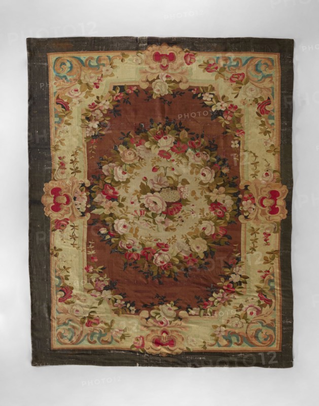 Ornamental tapestry, In the center, a bouquet of flowers surrounded by a floral wreath. Floral garlands surround the central field. Wide border with horn motifs, flowers in the corners, and floral shells. Colors: cream and light brown background; pink, red, blue, dark brown, sand, and green ornamentation. 18th/19th century, possibly France. Fabrics; height 381 cm, width 304 cm., A rectangular textile with a dark outer border and an inner decorative frame surrounds a central oval area filled with floral arrangements; the overall palette includes brown, beige, cream, pink, red, green, blue, and touches of black. The inner frame is lighter with floral sprays and ornate scroll motifs at the corners and midpoints, showing roses and leafy garlands in shades of pink, cream, and green against beige and cream backgrounds. The central oval field is a warm brown filled with a dense bouquet of flowers, featuring cream and pink roses, small red blooms, and green leaves with darker accents. The bord...