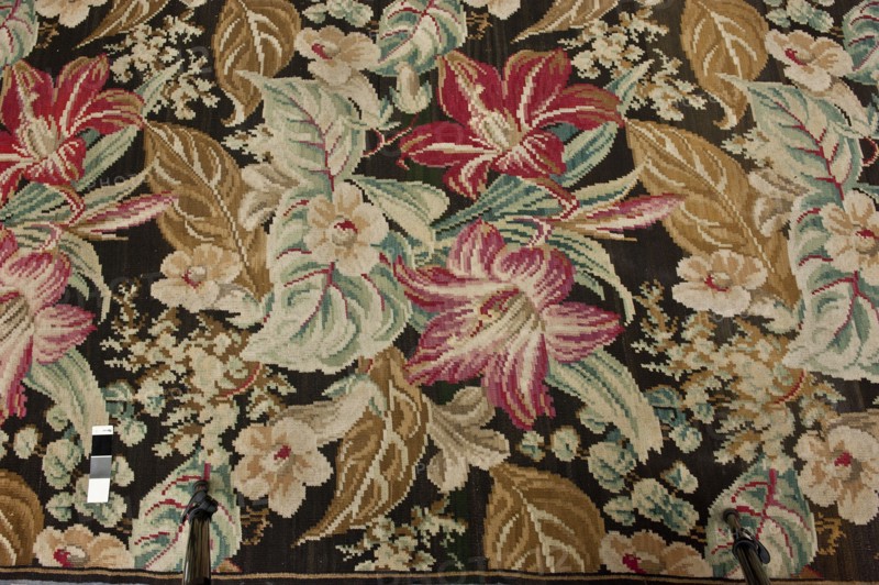 Kilim, Repossession provenance. Flowers; decorative fabrics; modernism style. Manor and home furnishings. Fabrics with plant and floral motifs; weaving. Kilim with motifs of large flowers and leaves filling the entire surface; no border. Colors: black background with light red, green, cream, pink, ash, and brown ornamentation. Late 19th century, Europe. Fabric kilim. Height 725 cm, width 450 cm., A woven textile with a dark background patterned with a floral design in shades of pink, red, cream, beige, tan, light blue, teal, green, and brown; the design includes large stylized blossoms, several broad leaves, and clusters of smaller flowers and buds; along the lower edge two metal tripod legs with black parts are visible and a small rectangular color strip with contrasting panels lies nearby.