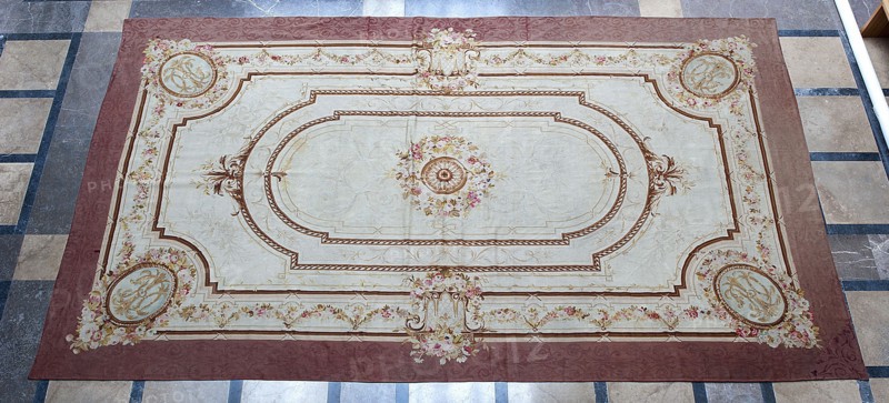 A so-called ceiling tapestry, tapestries, restitution provenance, fabrics, tapestries, knotted monograms, floral motifs, interior furnishings, weaving. In the center, a rosette surrounded by a floral wreath. Surrounded by rectangular frames with rounded shorter sides and floral garlands. In the corners are medallions with a knotted CKJ monogram. The border varies in color with plant patterns imitating the damask technique. Colors: pink, red, light brown, and olive. Early 19th century, France. Fabrics, height 681 cm, width 385 cm., A large rectangular decorative rug lies on a tiled floor. The rug has a wide border in a muted reddish-brown and a central field in a pale cream. Ornate rope-like and floral decorative bands in shades of brown and gold form nested oval and rectangular frames across the rug. Floral bouquets in soft pinks, light yellows, and muted greens appear at several points, including a central floral medallion with a detailed circular motif in brown and gold. Corner medal...