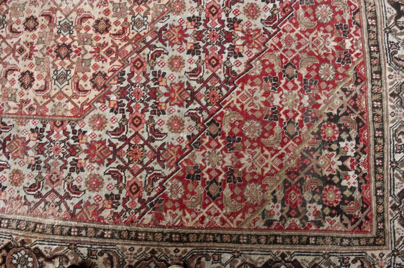 Carpet, Woolen knotted fabric, woven decorative carpet, 2nd half of the 19th century, Iran. In the center are four nested hexagonal medallions. Their interiors and the remaining background are filled with motifs of flowers, lanceolate leaves, and rosettes surrounding rhombuses. Three-band border with floral motifs. Colors: multiple tones of red, cream background, blue ornament. Height 667 cm, width 415 cm., A woven rug with an intricate geometric and floral pattern featuring a dominant mix of red and cream tones, accented by black, beige, brown, and hints of green. The design shows repeating medallions, star-like shapes, and small floral motifs arranged in diagonal bands and dense clusters, with a narrow ornamental border along one edge. The texture of the weave is visible, and the colors vary in intensity across the surface, creating areas that appear more saturated in red and others more muted in cream and beige. There is a subtle contrast between the darker black and brown accents a...