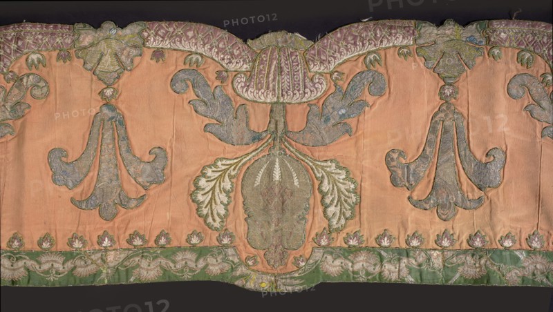 Lambrequin. Fragment of a lambrequin made of crimson silk grosgrain, sewn with fragments of patterned silk fabrics in green and brown. Decorated with palmette motifs, large serrated leaves, and small leaves arranged along the edges. The edges of the lambrequin are gently serrated. Silk lining. Early 18th century. Poland embroidery, France fabric. Materials: silk. Dimensions: height 41 cm, width 214 cm., Classification: decorative fabric / lambrequin., A decorative textile panel with a peach background featuring a central stylized floral motif in green and blue with pale green and white leaf shapes, flanked by two mirrored scrolling leaf designs in bluish-gray and green; a scalloped top border shows a pattern in purple and green, and a lower border band in green with pale green and white floral ornamentation; small stylized flower shapes in pink and white sit above the lower border; visible stitching and seams give a slightly wrinkled texture, and the overall palette includes peach, gre...
