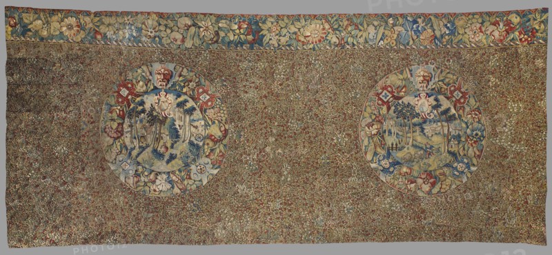 Tapestry, late 16th century, Flanders. Fabric fragment: entire background densely strewn with motifs of small flowering plants millefleurs. At the upper edge, two medallions with landscapes inside, their borders formed by floral and grotesque motifs; above them a border with motifs of flowers, fruit, and leaves. Decorative tapestry with flowers, fruit, medallions, and landscapes. Colors: black background, red, green, dark navy, and sand ornament. Materials: fabrics. Dimensions: height 167 cm, width 365 cm., A large rectangular textile with a dense floral field in muted green, brown, red, blue, cream, and gold tones, featuring two large circular medallions set within the patterned background; each medallion contains a scenic vignette in blue, cream, green, and beige hues surrounded by decorative floral borders in red, blue, cream, yellow, and green, and topped by a small mask-like face in cream and brown; a wide floral border runs along the top edge with stylized flowers and leaves in b...