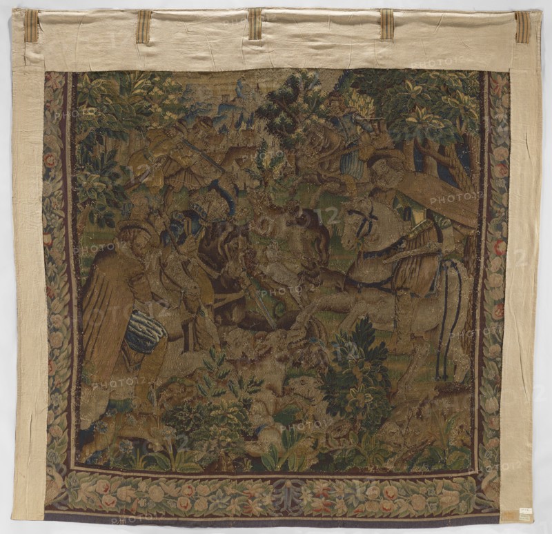 Figural tapestry, Scene: in the foreground at the edge of a forest, among low vegetation, dogs attack a wild boar; above, horseback riders with sabers or rifles; in the distance, urban buildings and a castle on a hill. Border with a motif of densely interwoven flowers and leaves. Motifs: flowers, floral ornaments, dogs, hunting, hunters, trees, horseman, forests, weapons., woven tapestry, decorative fabric; colors: brown, green, beige, red, gray. Date: 2nd half of the 17th century. Place of origin: Flanders. Dimensions: height 287 cm, width 291 cm., A rectangular woven tapestry with a wide beige fabric border and a central scene depicting a wooded outdoor setting in muted greens, browns, blues, and golds; within the scene a group of human figures and animals are arranged among dense foliage, trees, and flowering plants, with a few figures wearing garments in shades of brown and blue and a few animals showing beige and brown tones; the tapestry has a decorative floral border in green, p...