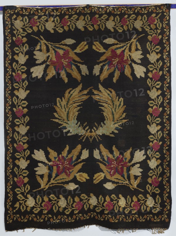 Kilim, In the center are motifs of two palm branches crossed at the base; in the corners are four branches with lily flowers. The kilim is surrounded by two floral tendril motifs. Colors: background dark brown; ornament variegated. Ca. 1850. Poland. Fabric. Height 201 cm; width 158 cm., A rectangular woven textile with a dark background featuring a central laurel-like wreath in shades of tan, gold, and muted green, surrounded by floral clusters in tan, gold, pink, red, and small accents of blue, with a decorative border of repeating floral motifs in similar tan, gold, pink, and red tones; the textile shows fringe at one short edge and the colors include black, tan, gold, pink, red, blue, and green.