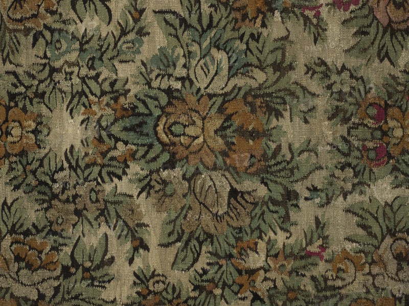 Court kilim. The main field is filled with flower and leaf motifs arranged symmetrically in repeating rhomboid arrangements. Surrounded by a three-stripe border: the middle, widest strip has a floral garland motif; the narrow outer strips feature floral and leafy tendrils. Colors: central field background off-white, borders tobacco-colored; ornamentation in green, magenta, tobacco, white, and black. Ca. 1850. Poland. Fabrics. Height 218 cm, width 215 cm., A patterned textile with an overall beige background featuring a repeating botanical motif in shades of green, muted teal, brown, tan, black, and small accents of magenta, showing stylized leaves and flowers in an ornate, symmetrical arrangement, with visible woven texture and areas of lighter and darker wear across the fabric.