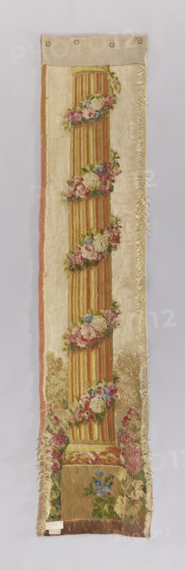 Ornamental tapestry, flowers, leaves, floral ornaments, columns, fabrics, interior decorations, interior furnishings, decorative fabrics. The column is identical to those flanking the pastoral scene in the tapestry inventory no. SZT 2211. Identical colors: cream, red, pink, green, and yellow. 1750–1780, France. Medium: fabrics. Height 225 cm, width 48 cm., A long vertical textile panel with a central fluted column motif in warm yellow and orange tones, wrapped at intervals with garlands of flowers in pink, red, blue, and green; the column sits on a square plinth in muted brown and gold hues surrounded by more floral clusters. The background fabric is a pale cream color with a narrow border on one side in faded pink and a fringed edge on the other side; the panel shows areas of wear and fraying along the edges and surface. At the top a plain linen or canvas strip in beige with round metal rings is attached, and the bottom edge shows a darker brown strip of fabric. Overall colors visible...