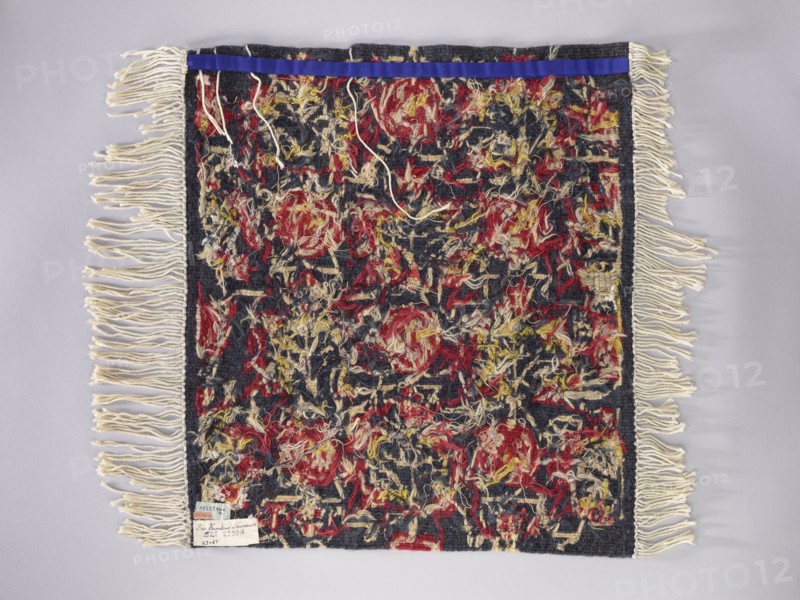 Tapestry covering; floral ornaments and stylized flower trees identical on the seat and backrest; decorative fabrics and borders; navy blue background with red, yellow, and cream ornamentation; fabrics; 1st half of the 20th century; Vienna; height 67 cm, width 74 cm., A square textile with a dense, multicolored central field framed by dark borders and long fringe on two opposite sides; the central area shows a textured, shaggy surface with a mix of red, yellow, cream, and dark blue fibers creating an irregular, mottled pattern, and a thin strip of bright blue along one edge; the fringe is cream colored and hangs loosely, while the background around the textile is pale gray.