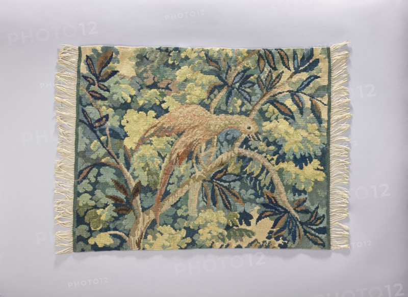 Tapestry chair cover, Repossession provenance. Design: leaves, floral ornaments, trees, birds, among thick foliage. Function: furniture upholstery/decorative fabric for interior furnishings. Materials: fabrics. Colors: yellow, green, blue, and pink. Date: 1st half of the 20th century. Place of origin: Vienna. Dimensions: height 65 cm, width 82 cm., A rectangular woven textile with fringe along the shorter edges lies flat against a pale gray background; the textile features a tapestry-like scene of plant forms and a perched bird, rendered in a palette of green, blue, yellow, brown, beige, and cream, with darker navy accents and touches of rust, showing leafy branches and layered foliage filling the composition.