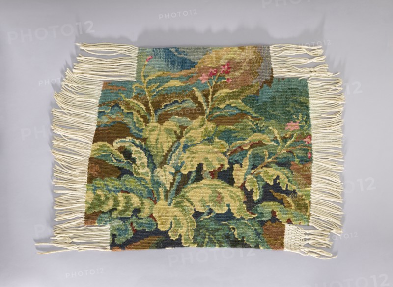 Tapestry chair cover, repossession provenance; design of flowers, leaves, and floral ornaments; decorative fabric for interior furnishings and upholstery gobelin-style tapestry; 1st half of the 20th century; Vienna place of origin; fabrics; height 68 cm, width 76 cm., A rectangular woven textile with cream-colored fringe along its shorter sides and parts of the longer sides, displaying a tapestry-like botanical design of large leafy forms and stems. The visible colors include various shades of green, teal, olive, brown, beige, blue, pink, and cream. The woven surface shows layered leaves with darker and lighter tones creating depth, and small areas of pink suggesting floral accents. The background areas transition between teal and brown tones, and the fringe is arranged in loose twisted strands against a light gray surface.