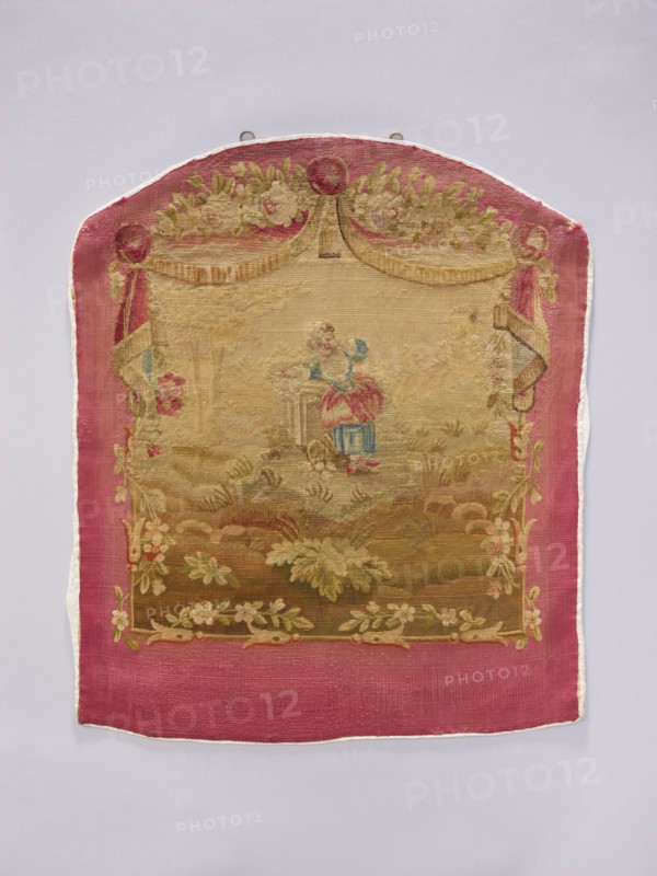 Tapestry covering, provenance, decorative tapestry with flowers, leaves, floral ornaments, girls, boys, and pastoral scenes; used for fabrics, interior decorations, furniture upholstery; design includes hanging draperies at the top and flowers around. Colors: beige, cream, green, sky blue, ochre, and red. Post–mid-18th century. Possibly Beauvais or Aubusson origin. Height 71 cm, width 58 cm., A rectangular textile with a rounded top hangs against a pale gray background; it has a wide pink border and a central woven scene in faded beige, tan, green, blue, and pink tones. The top of the textile features draped curtain-like shapes in beige and pink with floral garlands in green and pale pink, and a round decorative element at the apex. Within the central area a small figure wearing pink and blue sits near a pedestal or column, surrounded by a landscape of grassy and leafy forms in muted greens and browns. Floral garlands and leafy motifs frame the inner scene along the sides and bottom in...