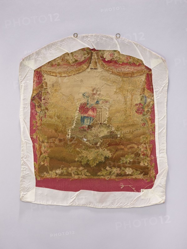 Tapestry covering, creator unknown, provenance, depicts flowers, leaves, floral ornaments, girls, pastoral scenes, boys; used as fabrics, interior decorations, interior furnishings, tapestries, furniture upholstery; with hanging draperies at the top and flowers around. Colors: beige, cream, green, sky blue, ochre, and red. Post–mid-18th century; possibly Beauvais or Aubusson origin. Height 71 cm, width 58 cm., A framed textile panel with a worn embroidered scene mounted on a pale background, showing a central figure in a dress with visible red and blue areas, surrounded by faded gold and beige embroidery and decorative floral motifs in shades of green and pink; the panel is bordered by a deep pink fabric and a white protective fabric wrap with metal hanging rings at the top, and the overall palette includes white, pink, red, blue, gold, beige, green, and brown.