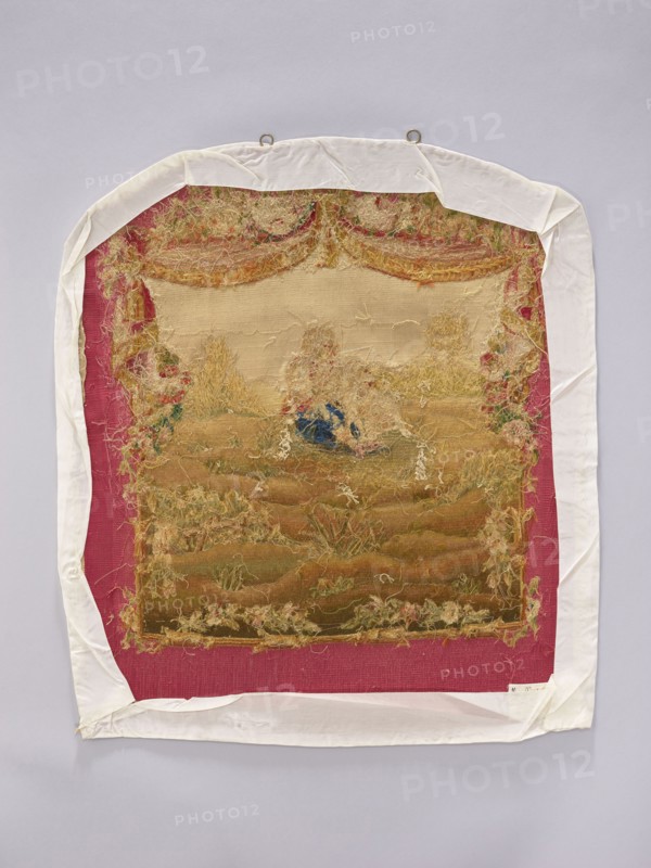 Tapestry covering, provenance, depicts flowers, floral ornaments, dogs, boys, pastoral scenes; used as interior furnishing, interior decoration, fabric for furniture upholstery; scene on grass with draperies suspended above and flowers around. Colors: beige, cream, green, ochre, blue, and red. Post–mid-18th century; possibly Aubusson or Beauvais. Fabric. Height 71 cm, width 59 cm., A rectangular textile with frayed edges is mounted on a pale background and wrapped with a white cloth border; the central fabric shows a worn tapestry scene in muted beige, tan, brown, and faded green tones with areas of loose threads in yellowish and cream hues and a small patch of dark blue; the tapestry is set against a deep pink or magenta backing visible around its edges; the white cloth border is slightly wrinkled and folded at the corners, and two metal loops are visible at the top.