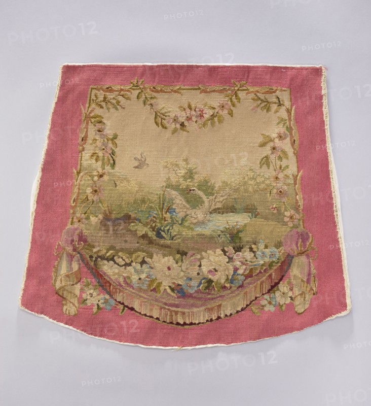 Tapestry covering, creator unknown, provenance, depicts flowers, leaves, floral ornaments, swans, boys, a swallow at the top, draperies below and flowers around; used for interior decorations, fabrics, furniture upholstery; style: gobelins/decorative fabric; colors: beige, green, blue, cream, pink; after the mid-18th century; possibly Aubusson or Beauvais origin; height 58 cm, width 67 cm., A decorative woven textile with a wide pink border surrounding a central beige field that contains a scenic woven image; the central scene shows a pale-colored swan on a pond amid green foliage and grasses with a small bird flying above, and the area is framed by garlands of leaves and flowers in shades of green, pink, and cream; the lower part of the design features a floral swag and draped fabric elements in muted pinks, creams, blues, and greens, with tassel-like fringe details and additional small flowers; the overall color palette includes pink, beige, cream, green, blue, and hints of brown.