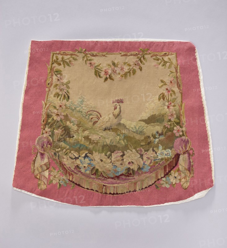 Tapestry covering, creator, flowers, leaves, floral ornaments, roosters, boys; decorative fabrics for interior furnishings and upholstery, draperies with flowers around the bottom. Colors: beige, light pink, sky blue, and olive green. After the mid-18th century. Possibly Beauvais or Aubusson origin. Height 56 cm, width 67 cm., A rectangular textile with a wide pink border and a central beige panel shows an ornate woven scene: a light-colored bird with a decorative crest stands amid a stylized landscape of green and brown hills and foliage, surrounded by floral garlands and leafy vine motifs in greens, pinks, and muted browns; the lower area features a draped swag of flowers and ribbons in shades of pink, purple, blue, cream, and green, with fringed tassel-like elements at the sides, and the outer edge trimmed with a narrow white border.