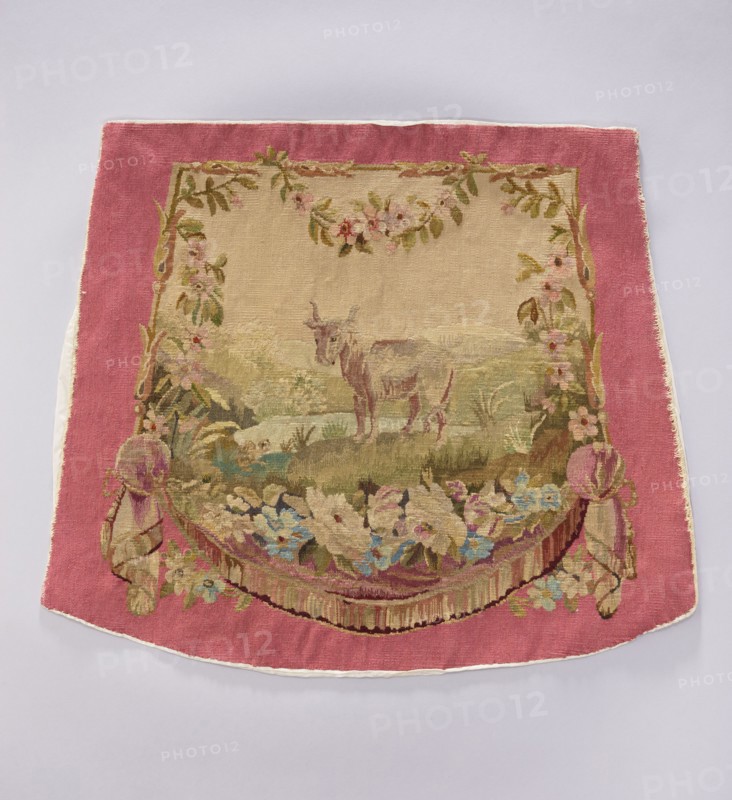 Tapestry covering, creator unknown, provenance, floral ornaments with flowers and leaves; oxen and boys; interior decorations and furnishings including tapestries, decorative fabrics, gobelins, furniture upholstery, and draperies with flowers at the bottom. Colors: beige, pink, sky blue, ochre, and olive. Post–mid-18th century; possibly Beauvais or Aubusson. Materials: fabrics. Dimensions: height 57 cm, width 68 cm., A rectangular woven textile with a wide pink border surrounds a central beige panel depicting a pastoral scene; the central area shows a light brown animal standing on grassy terrain with muted green and tan hills behind it, framed at the top and sides by garlands of leaves and flowers in shades of green, pink, and pale yellow. The lower edge of the central scene features a curved floral swag with a mix of soft blue, cream, pink, and green flowers and foliage, and draped tassel-like elements in pink and beige at the lower corners. The outer border is solid rose-pink, and t...