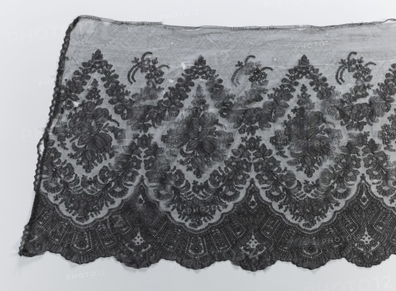 Crinoline ruffle, clothing accessories, garlands ornament, Chantilly lace, floral ornaments, lace, fashion. Bouquets placed in medallions created with garlands above and below them. Below the lower garland, a fan-shaped lace motif. The lower edge is serrated, the upper one straight. 2nd half of the 19th century, Chantilly place of origin, clothing, lace, width 54 cm, length 440 cm, A section of patterned lace fabric laid flat, showing an intricate floral and scalloped design in dark gray and lighter gray tones; the lace has a sheer background with denser embroidered floral motifs and swirling leaf shapes, scalloped edges along the bottom with repeating fan-like arches, and a slightly uneven top edge; a few loose threads are visible along the sides, and the overall color palette is shades of gray against a pale gray background.