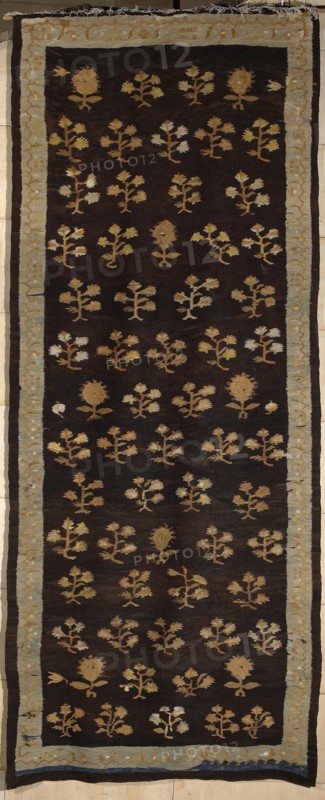 Kilim, kilim, flowers, interior design, Poland culture, decorative fabrics, floral patterns. The main field of the kilim is filled with motifs of stylized flower branches arranged in alternating vertical rows; the branches are four-flowered or single-flowered. Surrounded by a border with a geometrical floral tendril motif. Colors: background of the main field is brown, borders are light blue, ornamentation golden yellow. ca. 1830, Poland, fabrics, height 376 cm, width 148 cm., A long rectangular textile with a dark brown background framed by a pale blue-gray border; the interior is patterned with a repeating arrangement of small stylized floral motifs in shades of tan, beige, light brown, and muted yellow, with a few motifs showing lighter off-white highlights; the border contains a subtle decorative motif in similar pale tones and the outer edge shows narrow bands of darker brown and a thin strip of deep blue; the textile shows frayed edges at one end and slight irregularities in weav...